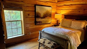 A wooden cabin bedroom with a bed, bench, window, and painting.