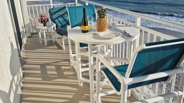Un balcon avec vue sur l’océan, agrémenté de chaises blanches, d’une table avec une bouteille et d’un ananas.