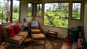 A cozy living room with a sofa, armchair, and a small table. Large windows offer a view of lush greenery outside.