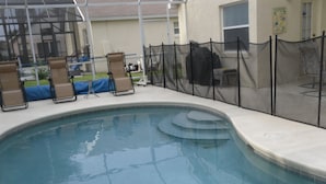 Une piscine à bord incurvé, entourée d’une clôture en treillis noir. On y trouve des chaises longues avec des housses bleues et, au fond, un barbecue.