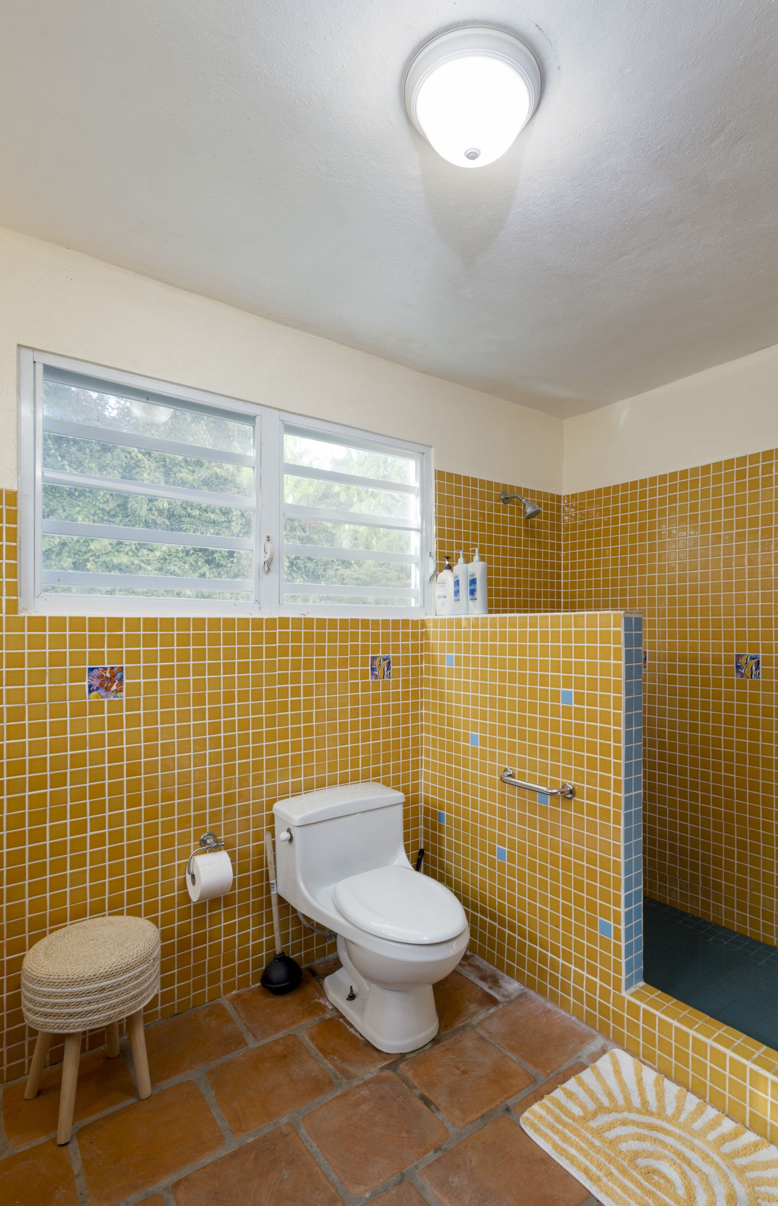 Une salle de bain avec des murs recouverts de carreaux jaunes, des toilettes blanches, un petit tabouret, une fenêtre et un espace douche.