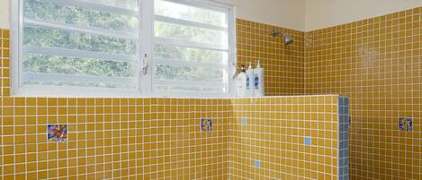 Un baño con paredes de azulejos amarillos, inodoro blanco, un taburete pequeño, una ventana y una zona de ducha.