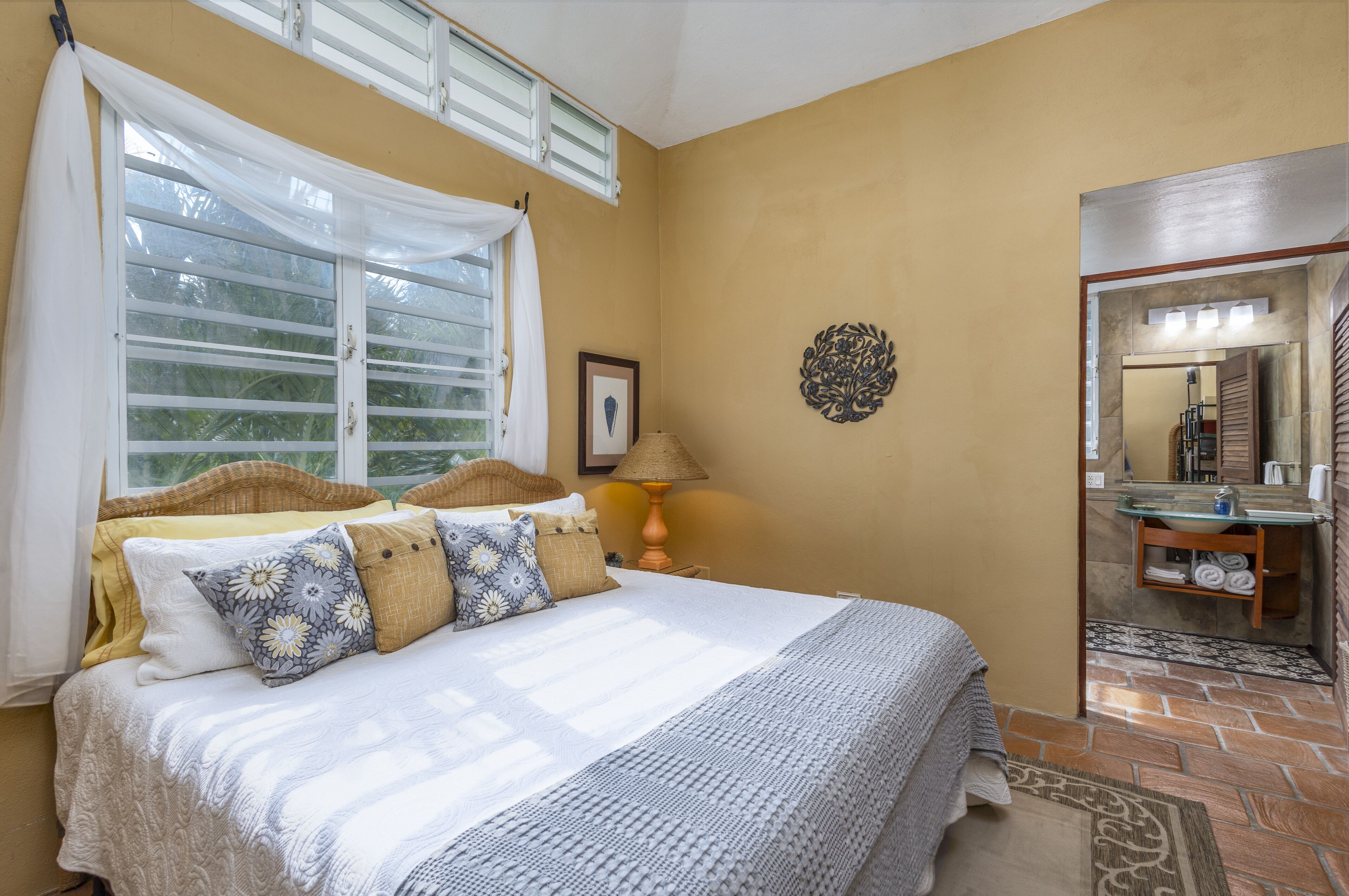 A bedroom with a bed, a window with curtains, a bedside lamp, a framed picture on the wall, and a bathroom visible through an open door.