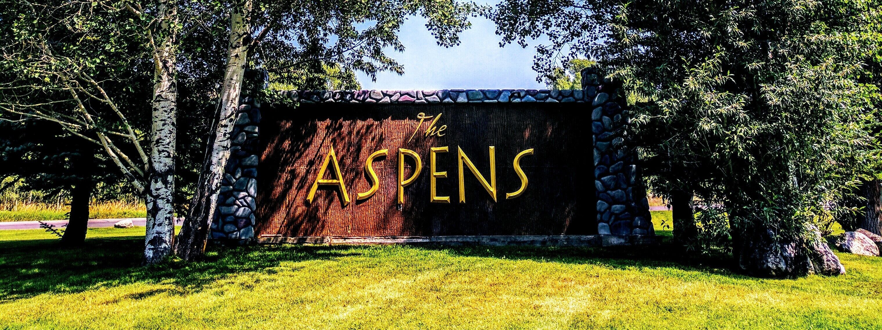 A wooden sign with the name "The ASPENS" in gold lettering, surrounded by trees and a stone wall.
