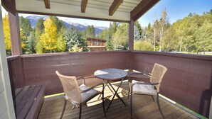 Une terrasse extérieure couverte, avec une table en bois et deux chaises en osier, donnant sur une forêt et des montagnes.