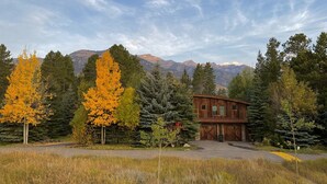 Un chalet rustique aux murs en bois, entouré d’une forêt aux couleurs automnales, avec des montagnes en arrière-plan.