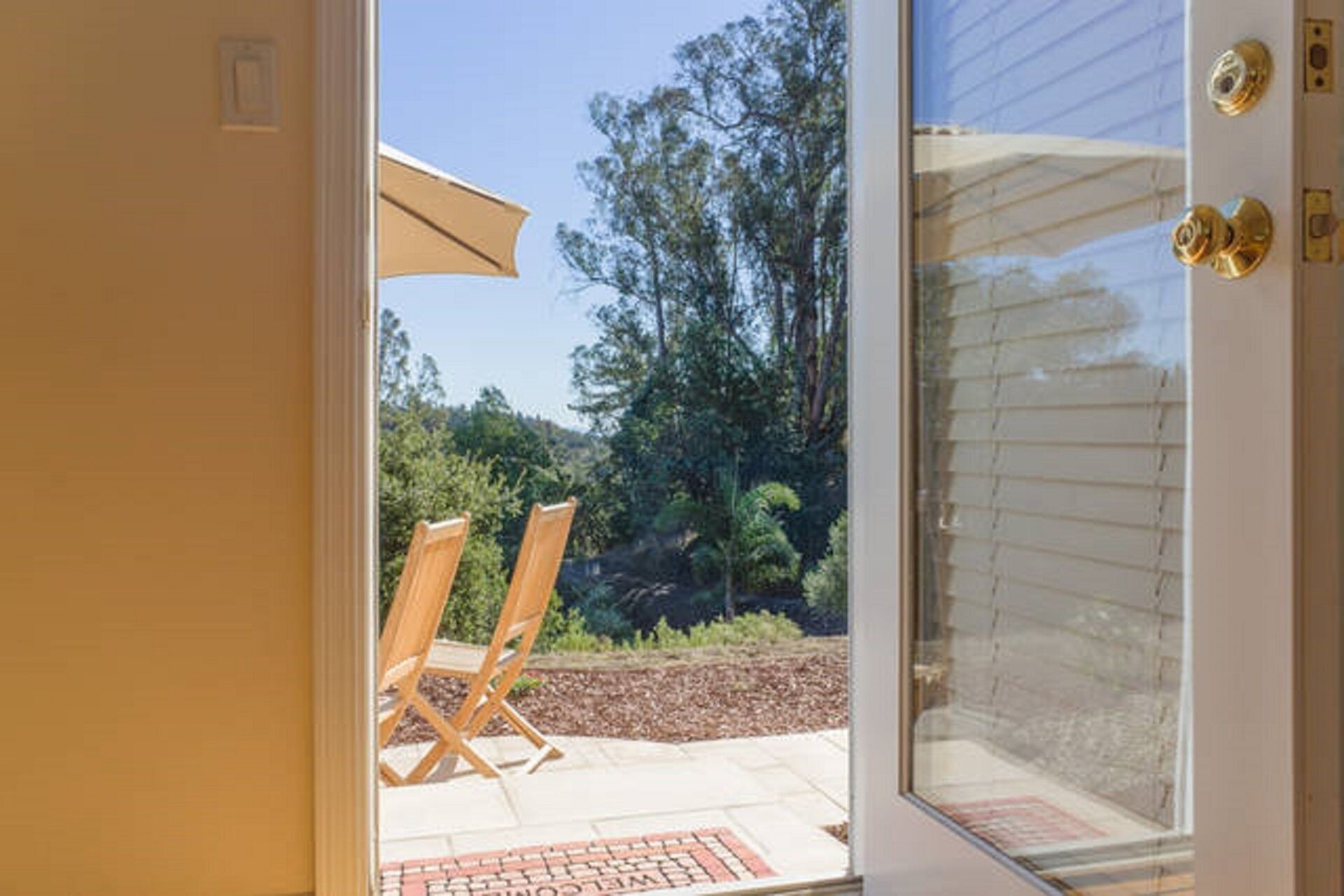 A glass door leading to an outdoor patio with two wooden chairs and a table, surrounded by trees and a clear sky.