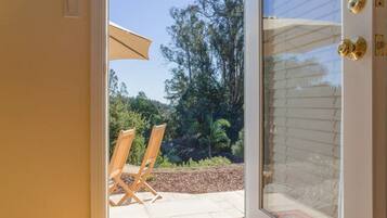 A glass door leading to an outdoor patio with two wooden chairs and a table, surrounded by trees and a clear sky.
