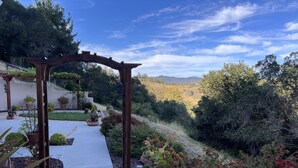 Ein Gartenpfad führt zu einer hölzernen Pergola mit Blumentöpfen und Blick auf eine hügelige Landschaft.