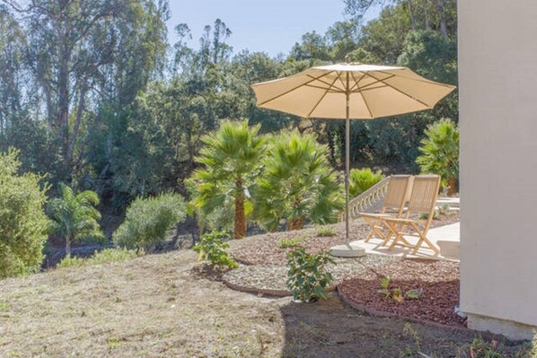 A patio area with a beige umbrella, wooden chairs, and a garden with various plants and trees.