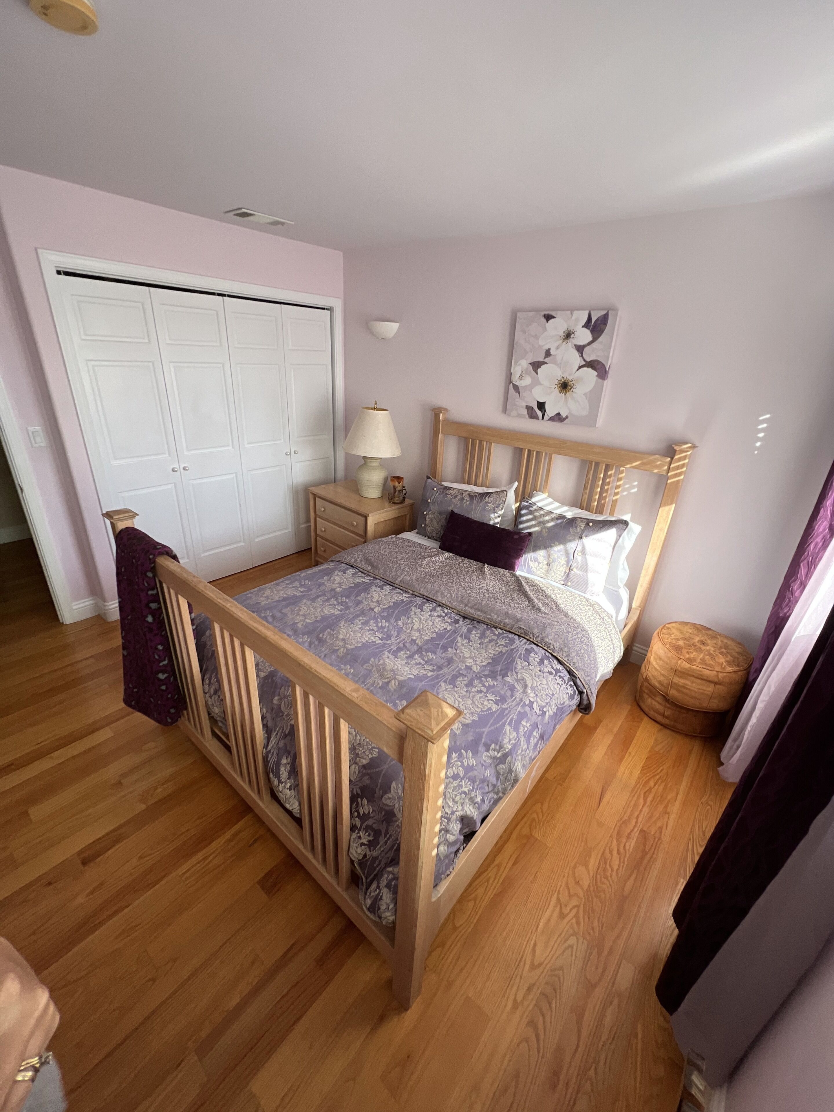Ein Schlafzimmer mit einem hölzernen Bett, einem Nachttisch, einer Lampe, einem Kleiderschrank und einer Wand mit einem floralen Gemälde.