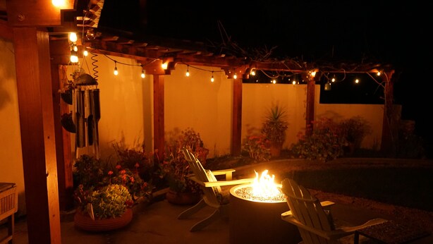 Eine Außenpatio-Terrasse mit Feuerstelle, Sitzgelegenheiten, Lichterketten und Topfpflanzen.