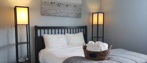 A neatly made bed with white linens, two standing floor lamps, and a wall-mounted artwork depicting a beach scene.