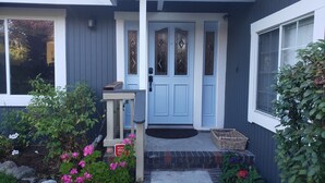 Et hus med en blå dør, en lille veranda og en have med lyserøde blomster.