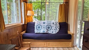 Interior acolhedor de uma cabana com um banco de madeira, um tapete estampado e vista para o verde lá fora.