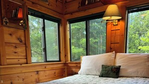 Quarto de cabana de madeira com cama, travesseiros, luminária e janela com vista para uma floresta.