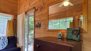 Interior de cabana com teto de madeira, cama com colcha, cómoda, espelho e janela com cortinas.