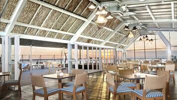 Un vaste espace extérieur aménagé pour les repas, avec des tables et des chaises en bois, de grandes fenêtres offrant une vue sur le coucher de soleil, et un plafond haut avec des poutres apparentes.
