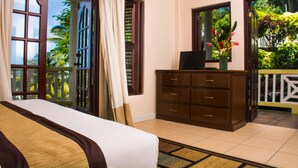 Ein Hotelzimmer mit Bett, Schminktisch, Schreibtisch mit Computer, Fenster mit Blick auf Grünflächen und einer Tür, die zu einem Außenbereich führt.