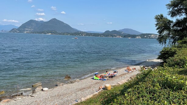 Una playa de guijarros con gente tomando el sol, un barco en el agua y montañas al fondo.