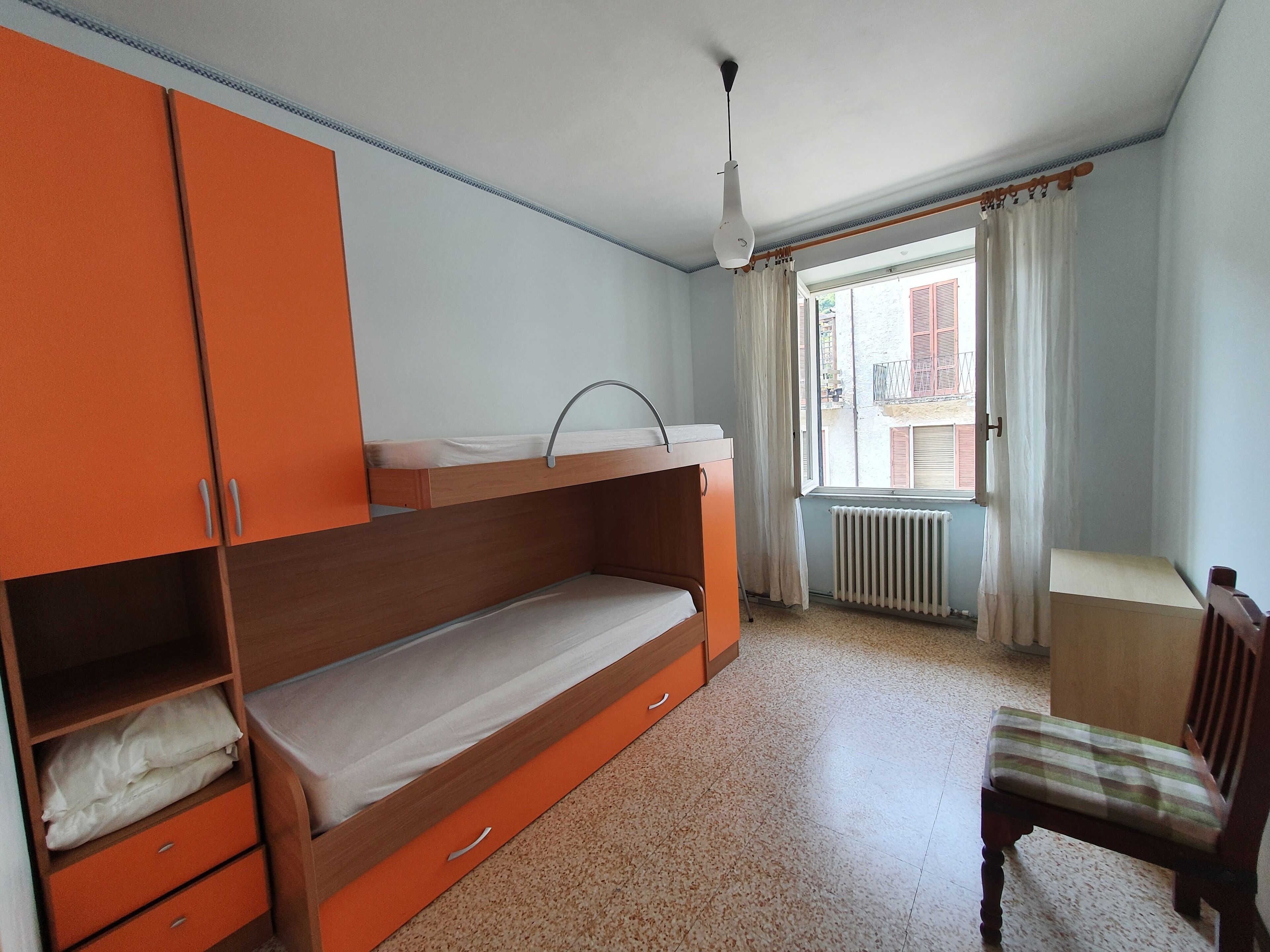 A room with a bunk bed, a wooden wardrobe, a radiator, a window with curtains, and a chair.