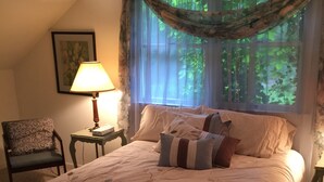 Ein Schlafzimmer mit Bett, Sessel, Lampe und Fenster mit Blick auf Grünfläche.