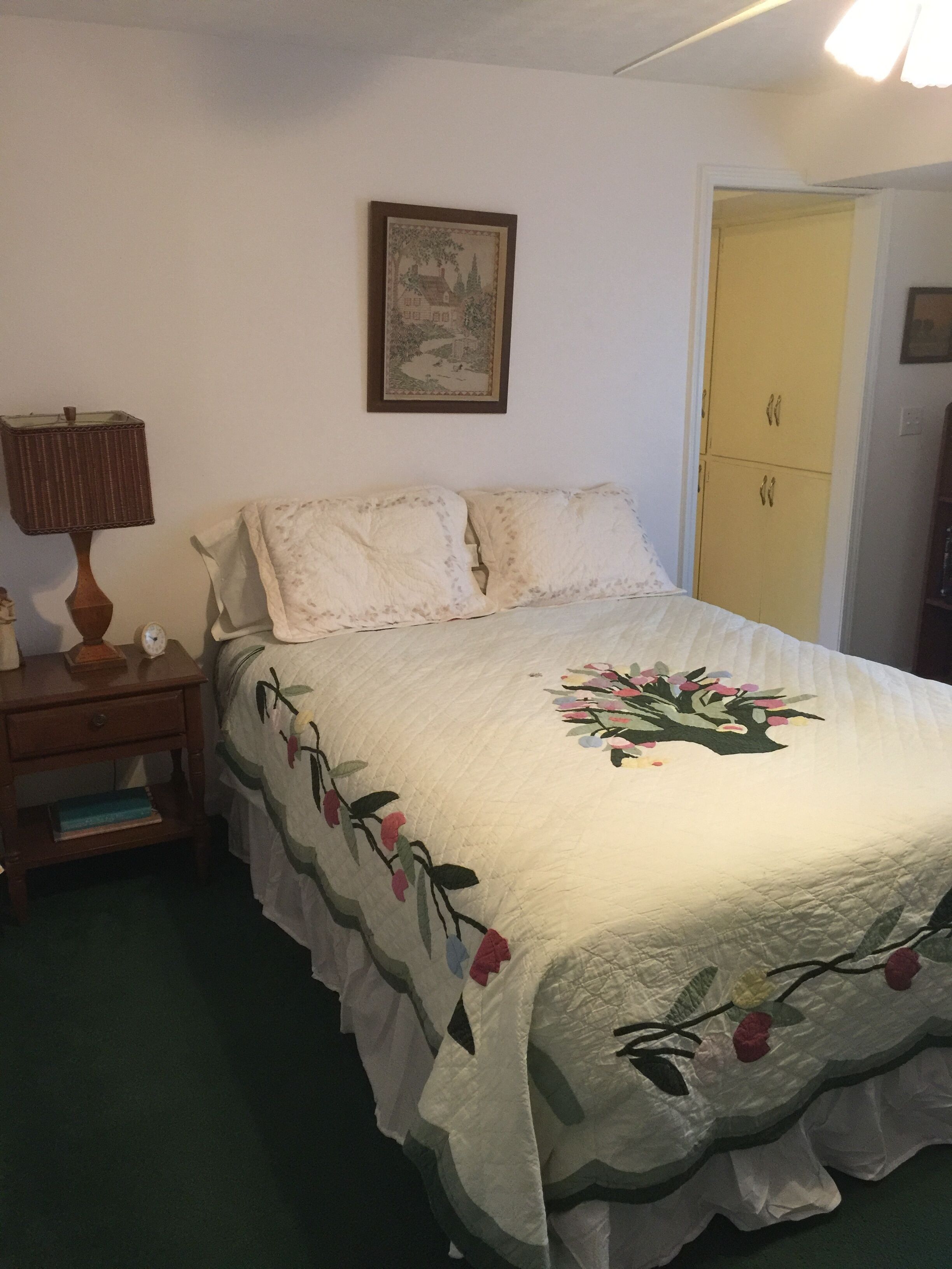 A bedroom with a bed, a nightstand, a lamp, a clock, a framed picture on the wall, and a yellow door.
