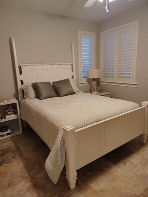 A bedroom with a white four-poster bed, a nightstand with a lamp, a small shelf with books, and a window with blinds. - Luxurious, peaceful beachfront condo minutes from St Augustine and Daytona Beach (Beverly Beach)