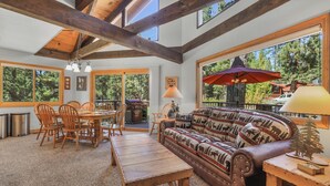 Amplia sala de estar con mesa de comedor de madera, sillas y sofá con cojines con motivos de animales. Grandes ventanas ofrecen vistas al exterior.