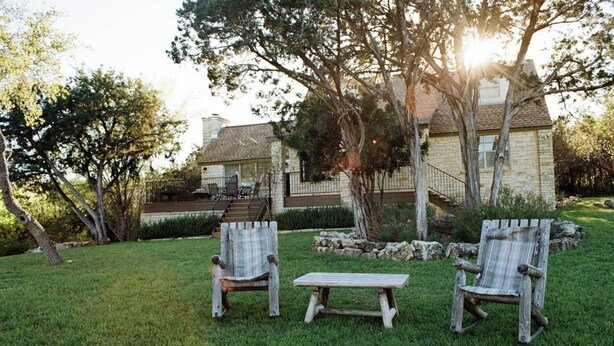 A stone house with a covered patio, two wooden chairs, and a small wooden table in a grassy yard. - 4/2 by Marina, Near Wineries, Grill, WiFi, View (Spicewood)