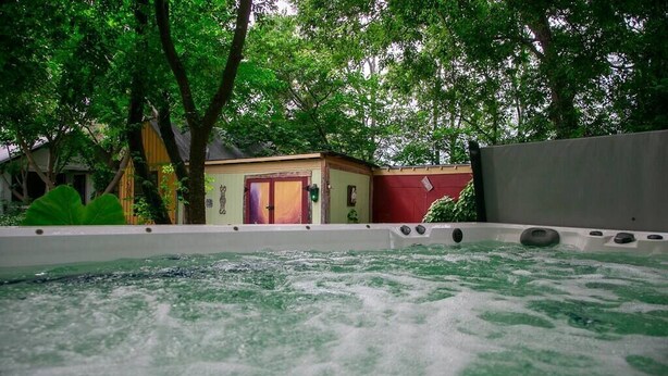 Een hottubgedeelte met een houten terras, omringd door bomen en een klein gebouw.