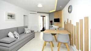 Un salon moderne avec un canapé gris, une table à manger en bois accompagnée de deux chaises, un téléviseur à écran plat fixé au mur et une grande fenêtre avec des rideaux.