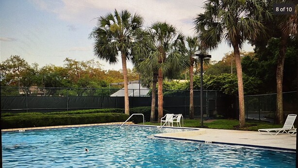 Una piscina con palmeras, un área cercada y una estructura cubierta al fondo.