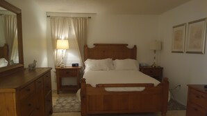 Un dormitorio con cama de madera, una mesita de noche, cómoda, espejo y ventana con cortinas.