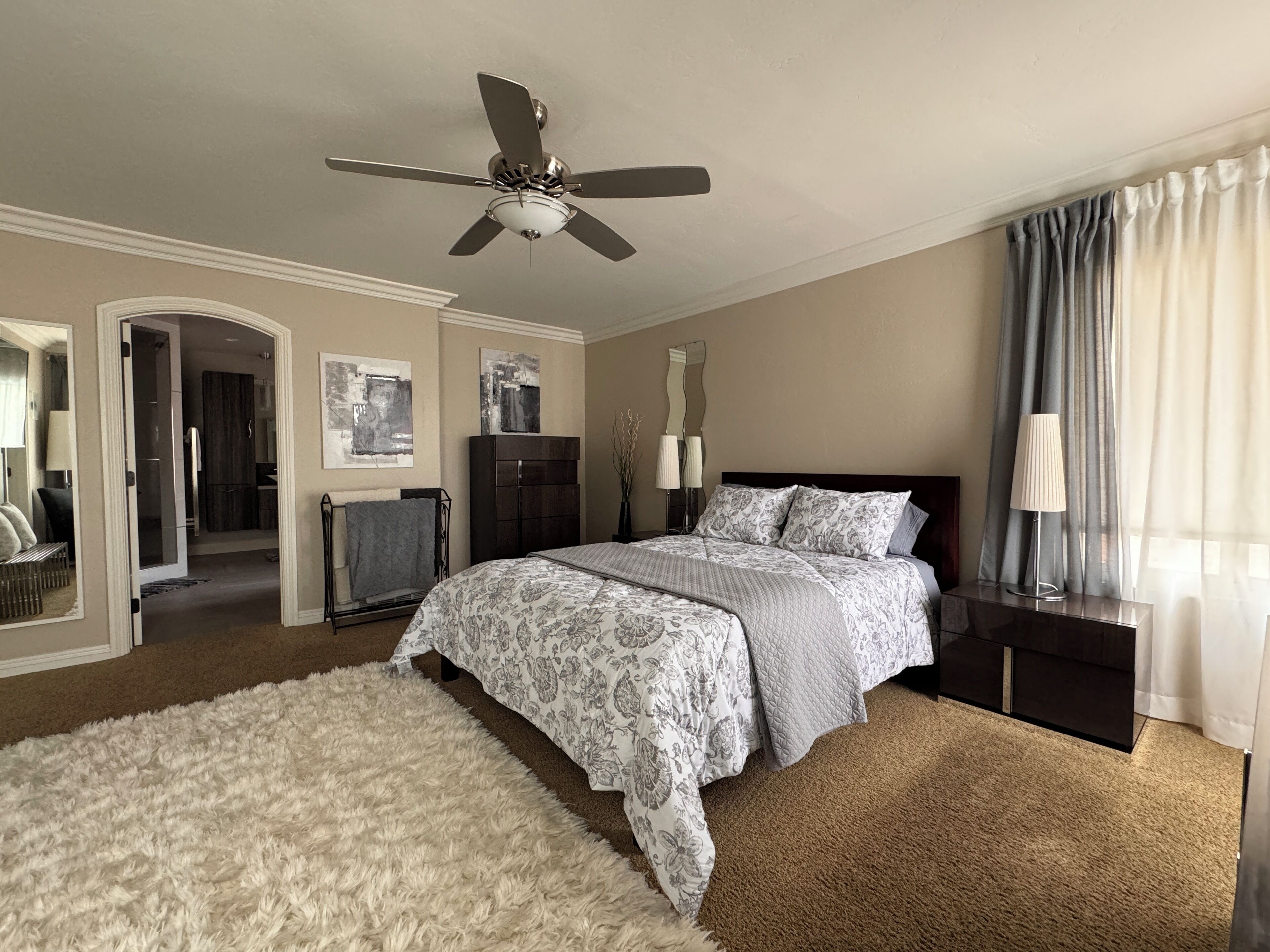 Une chambre à coucher comprenant un lit, un ventilateur de plafond, une table de chevet, une lampe et un grand miroir.