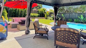 Ein überdachter Patio-Bereich mit Gartenmöbeln, darunter Korbstühle und eine gepolsterte Bank, unter einem Metalldach mit roten Sonnenschirmen.