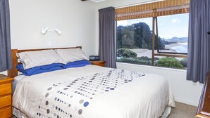 A bedroom with a large bed, a bedside table, a nightstand, and a window with a view of the beach. - Fantastic Family Holiday Home with Breathtaking Views over Hot Water Beach (Hot Water Beach)