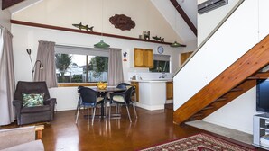 A spacious living area with a dining table, chairs, and a comfortable armchair. A kitchen area is visible in the background with a window and a clock. - Fantastic Family Holiday Home with Breathtaking Views over Hot Water Beach (Hot Water Beach)