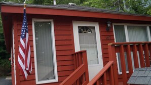 A red wooden cabin with a white door, a window, and an American flag.