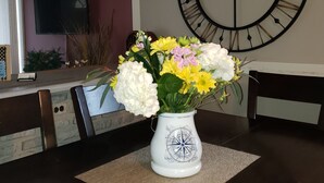 A dining table with a white vase of flowers, a large wall clock, and a television mounted on the wall.