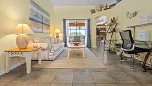 A living room with a sofa, two lamps, a coffee table, and a desk with a chair. There is a large painting on the wall and a window with curtains. - VERY CLEAN & SPACIOUS! Pool, Game Room, BBQ, 2 King Masters--Close to Disney! (Clermont)