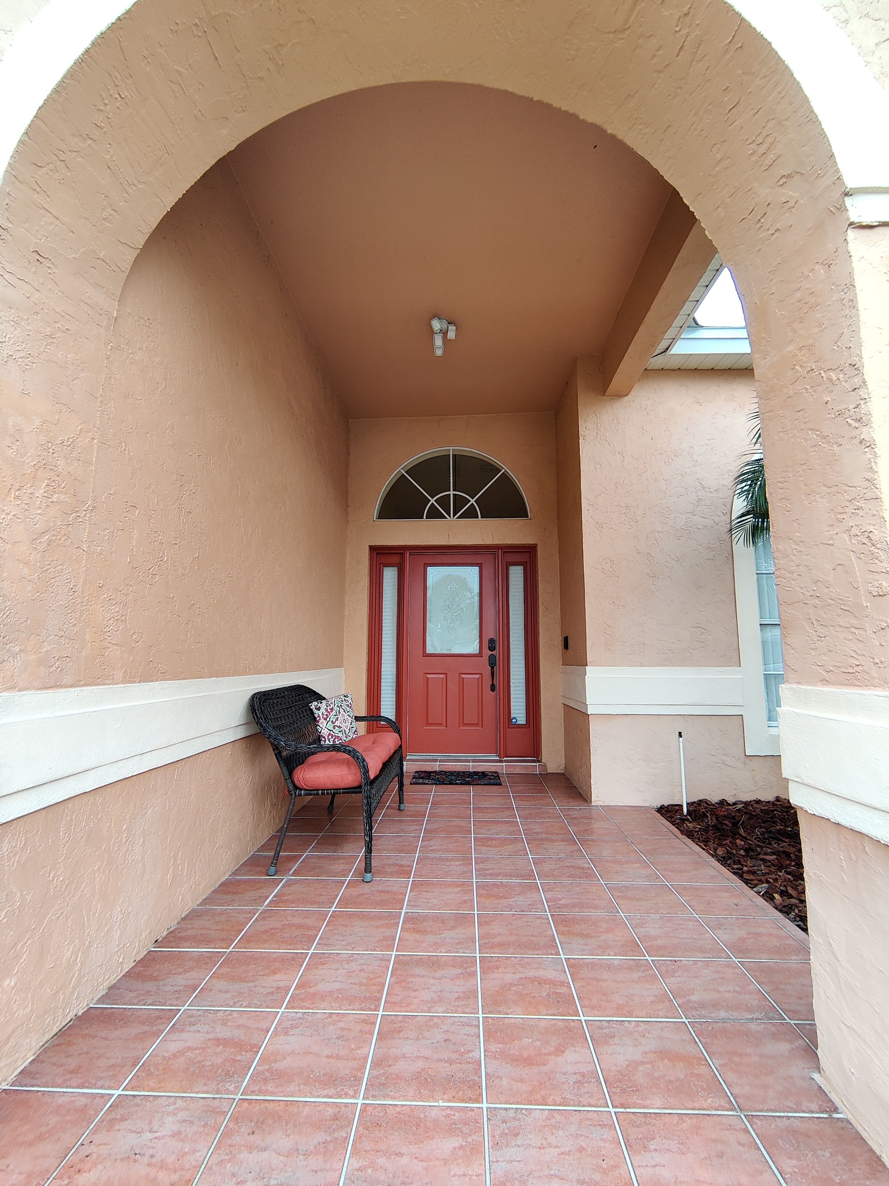 A covered entrance with a red door, a wicker chair with a cushion, and a tiled floor.