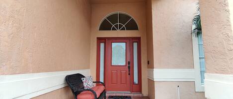 A covered entrance with a red door, a wicker chair with a cushion, and a tiled floor.