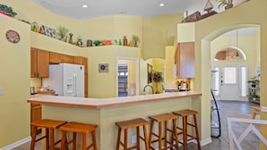 A kitchen with wooden cabinets, a white refrigerator, and a bar area with stools. - VERY CLEAN & SPACIOUS! Pool, Game Room, BBQ, 2 King Masters--Close to Disney! (Clermont)