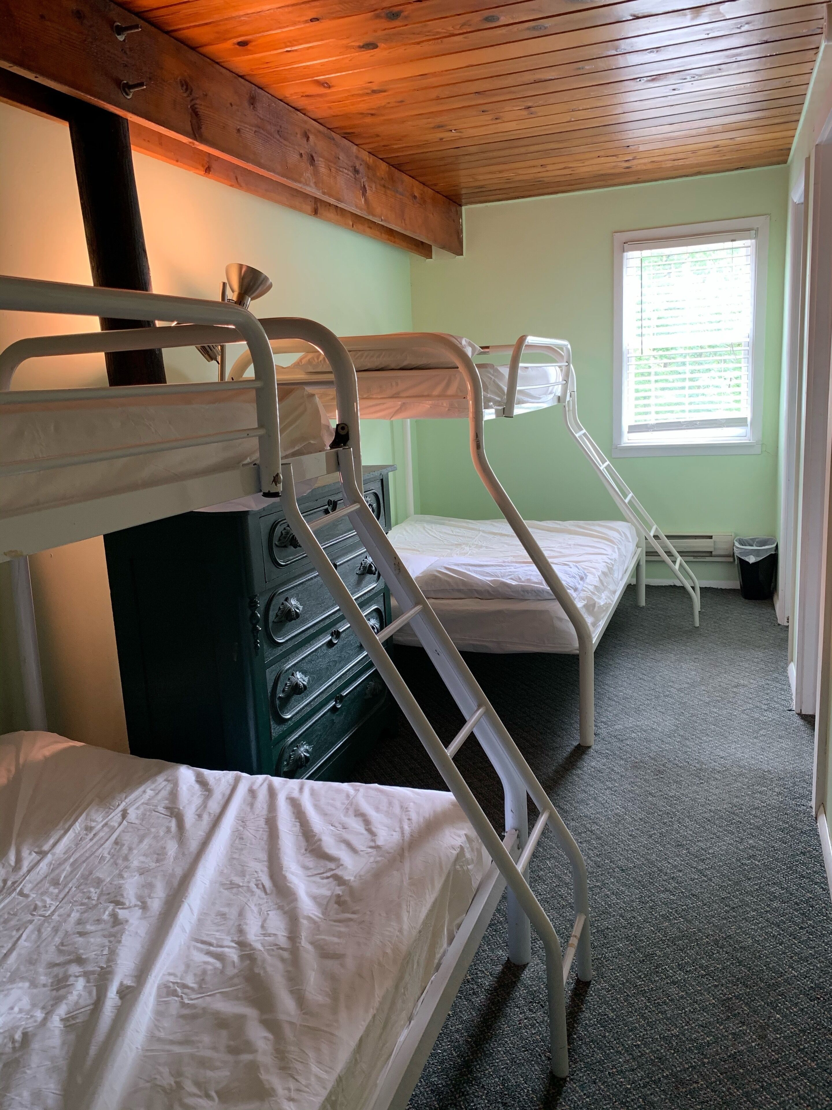 A bunk bed room with a wooden ceiling, a window with blinds, and a green dresser.