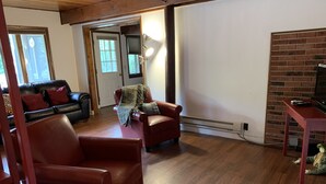 Een woonkamer met een rode leren bank, een rode fauteuil en een kleine tafel met een afstandsbediening. Er is een open haard en een raam met zonwering.