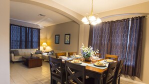 Un comedor con una mesa de madera dispuesta para cuatro personas, rodeada de sillas de madera oscura. En la zona de estar contigua, hay un sofá y una mesa de centro.