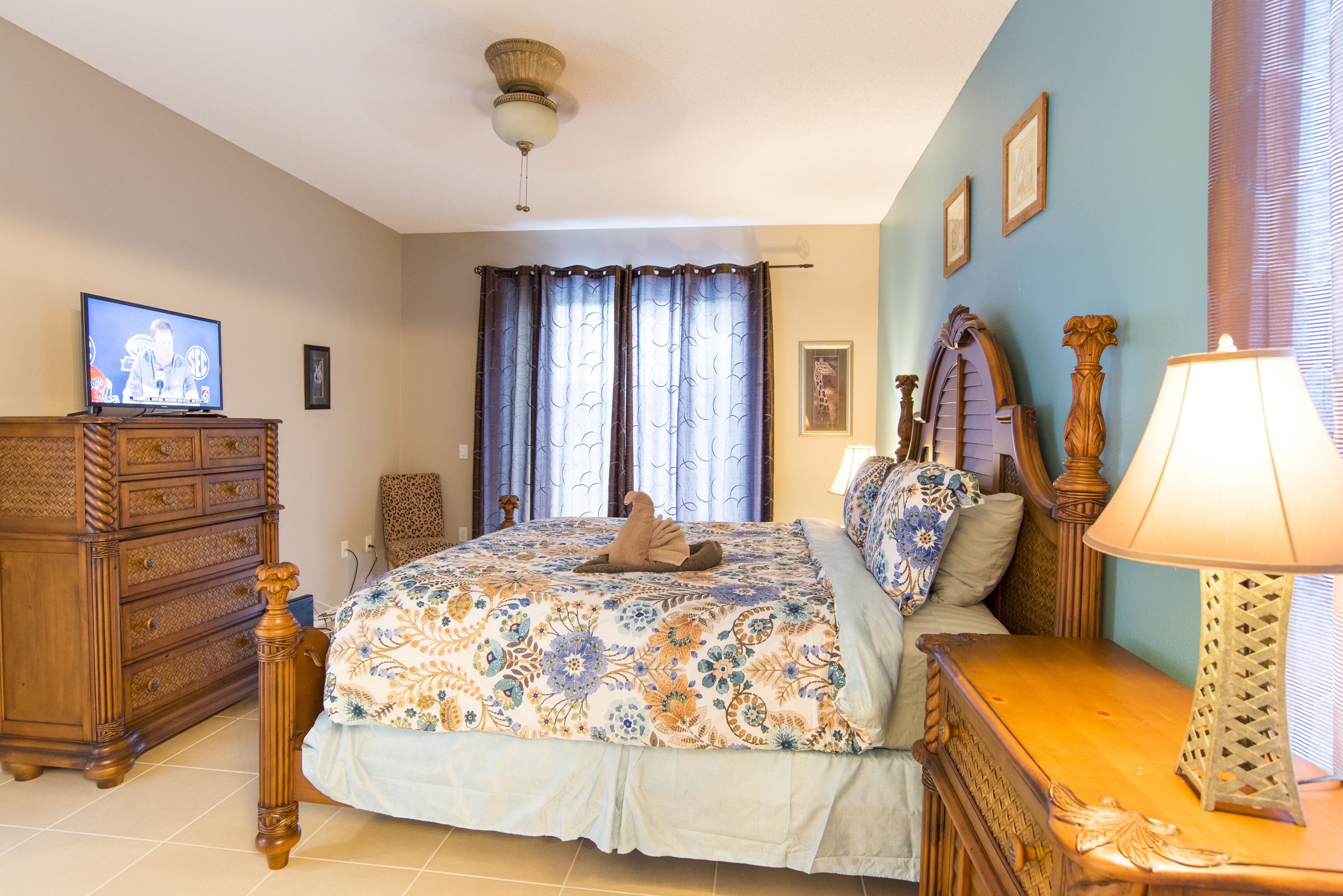 A bedroom with a bed, dresser, lamp, curtains, and a television.