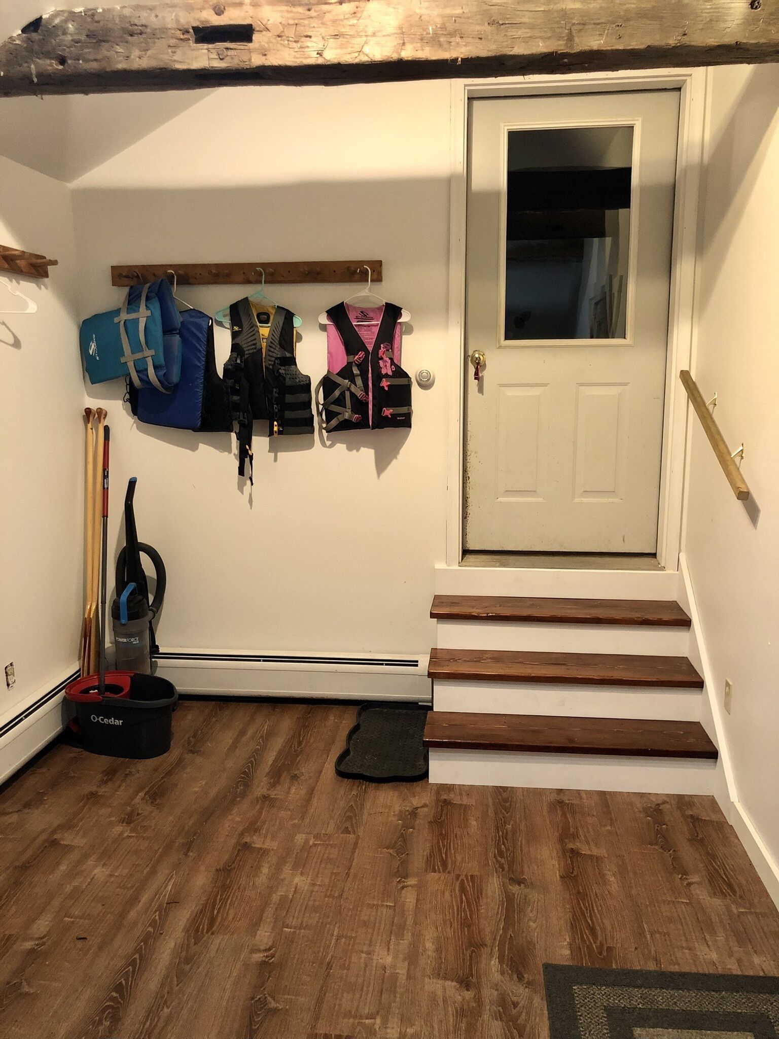 Una habitación con piso de madera, una puerta, un juego de escalones y almacenamiento en la pared para maletas y un aspirador.