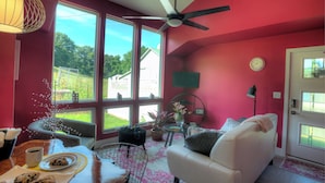 A cozy living room with a white sofa, a dining table with a coffee cup and cookies, large windows, a ceiling fan, and a view of a house and greenery outside. -  Modern country cottage that is dog friendly with a fenced-in yard.  (Columbia)